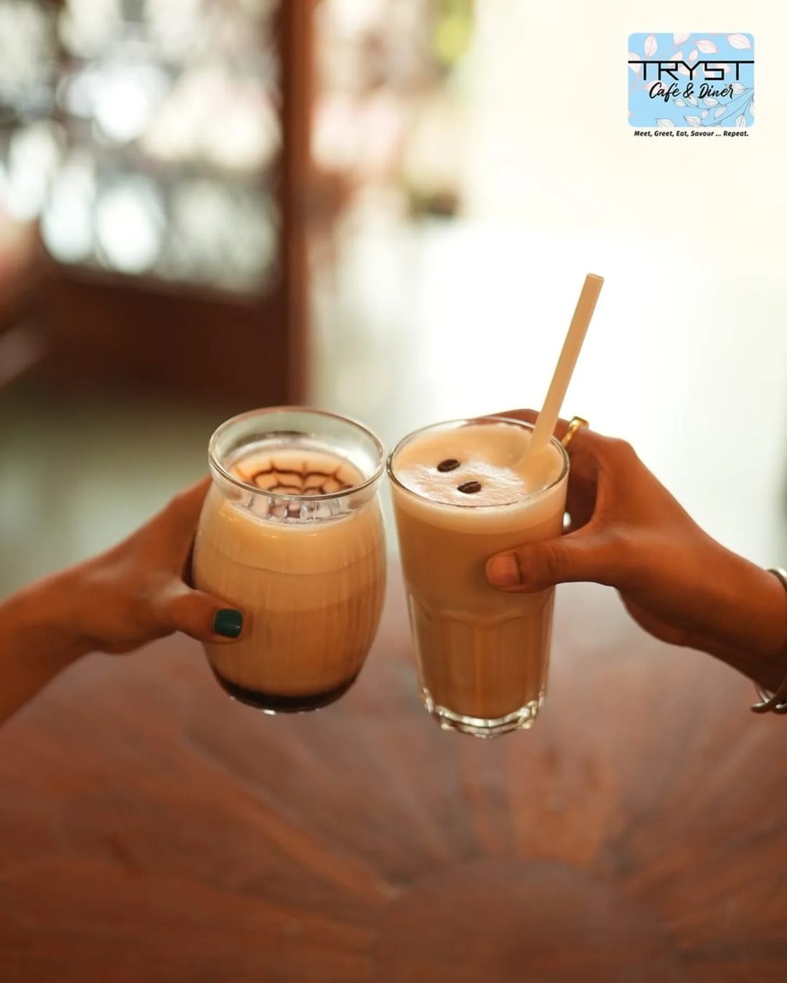 Two hands raising an iced mocha and an iced latte across a wooden table